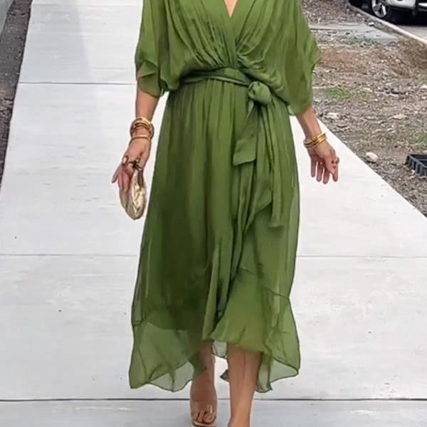 Femme portant une robe longue en chiffon vert olive, cintrée à la taille avec un nœud, et accompagnée d'un sac à main doré et des bracelets dorés.