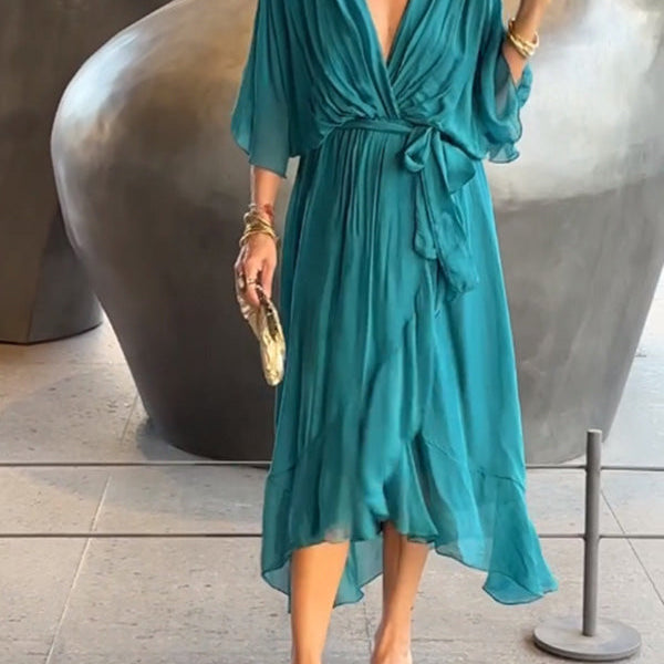 Femme portant une robe chiffon turquoise fluide avec un décolleté en V et des manches larges. Elle accessoirise la tenue avec une pochette dorée et des bracelets dorés.