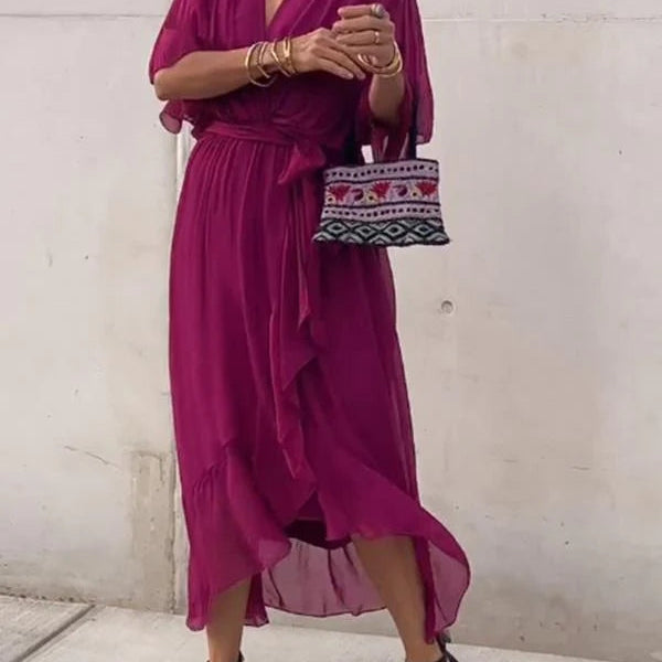 Femme portant une robe chiffon fuchsia avec des manches fluides et un décolleté en V. Elle accessoirise la tenue avec un sac à main tissé et des sandales noires à talons.