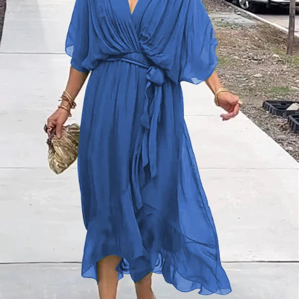 Femme portant une robe chiffon bleu fluide avec des manches larges et un décolleté en V, accessoirisée d'une pochette dorée et des bracelets dorés.