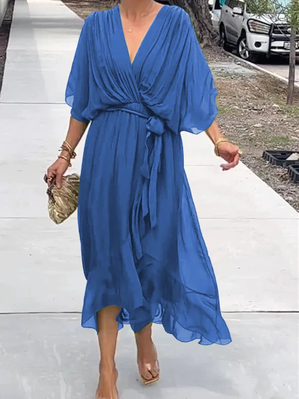 Femme portant une robe chiffon bleu fluide avec des manches larges et un décolleté en V, accessoirisée d'une pochette dorée et des bracelets dorés.