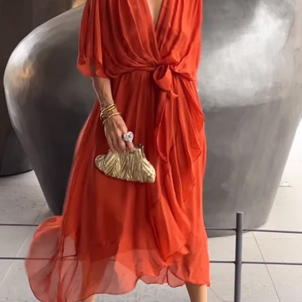 Femme portant une robe chiffon rouge vif, avec des manches amples et un décolleté en V, accessoirisée d'une pochette dorée et des bracelets dorés.