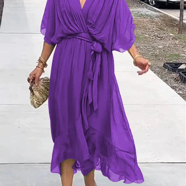 Femme marchant dans une robe chiffon violette, avec des manches larges et une ceinture assortie, portant une pochette dorée et des sandales transparentes.