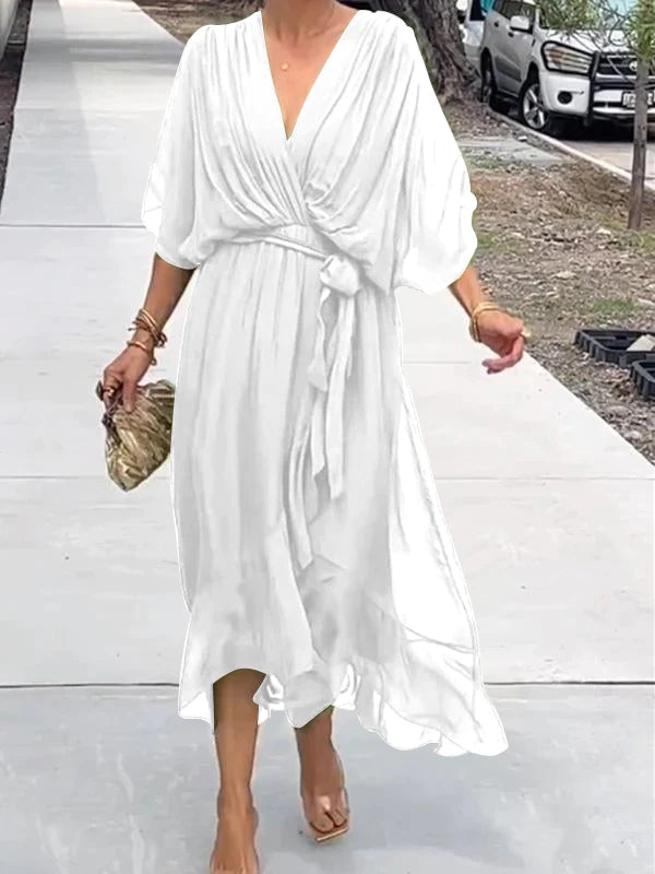 Femme marchant dans une robe chiffon blanche, avec des manches larges et une ceinture assortie, portant une pochette dorée et des sandales transparentes.