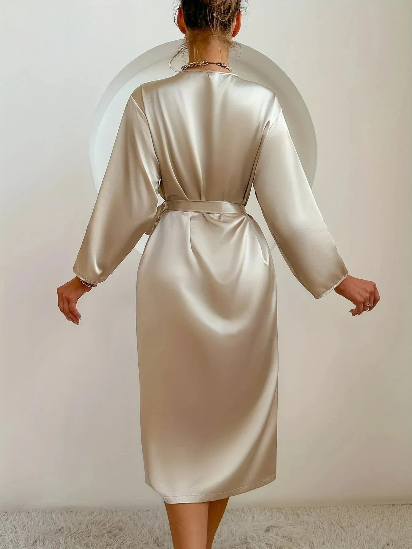 Vue de dos d’une femme portant un peignoir satiné couleur champagne, à manches longues et ceinture nouée à la taille. Modèle fluide, sobre et élégant, idéal pour une tenue de détente chic et confortable à la maison.