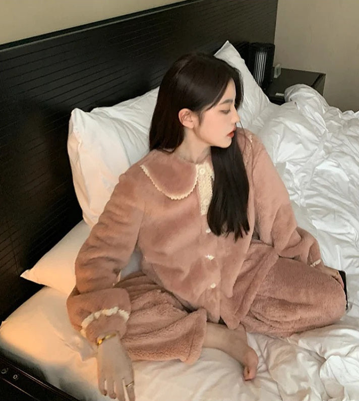 Femme assise sur un lit en pyjama polaire rose doux, avec col Claudine orné de dentelle crème et poignets assortis. Ensemble boutonné, chaud et élégant, parfait pour se détendre confortablement à la maison pendant l’hiver.