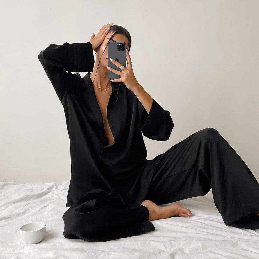 Pyjama femme Armelle noir, ensemble deux pièces raffiné et confortable avec chemise fluide décolletée à manches longues et pantalon large assorti, porté en intérieur sur un lit, tissu satiné élégant.
