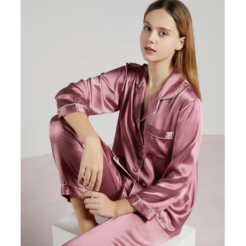 Femme assise portant un pyjama en satin vieux rose, coupe classique avec col à revers, manches longues et passepoils contrastants. Ensemble doux, brillant et raffiné, parfait pour les nuits reposantes ou les moments de détente élégants à la maison.