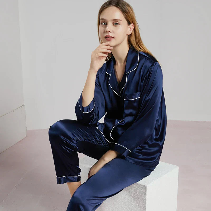 Femme assise portant un pyjama en satin bleu marine à manches longues, col à revers et finitions passepoilées blanches. Ensemble chic et confortable, idéal pour la nuit ou les moments de détente élégants à la maison.
