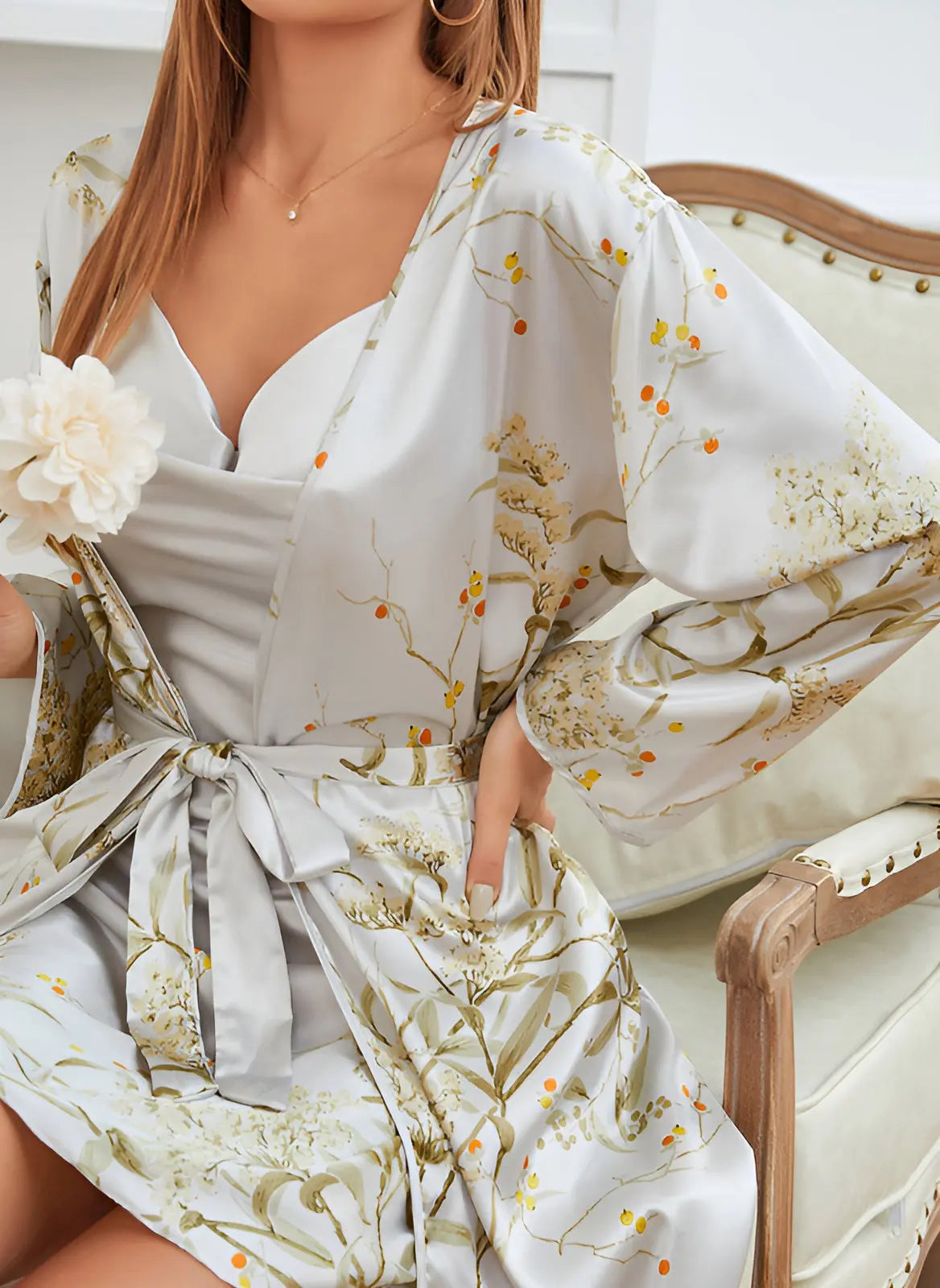 Détail de la robe-kimono satinée gris clair avec des motifs floraux délicats, ceinturée à la taille avec un nœud satin. La femme tient une fleur blanche, ajoutant une touche de douceur et d'élégance. Robe fluide et raffinée, parfaite pour des moments de détente ou une soirée chic.