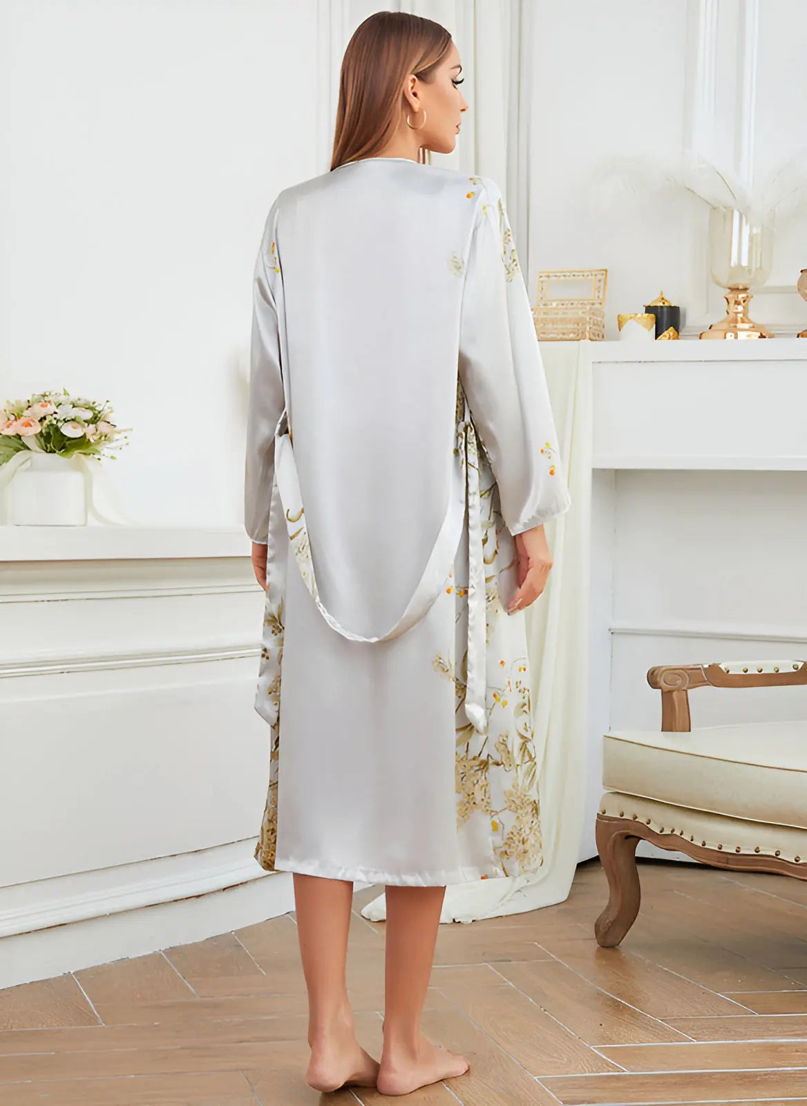 Vue arrière de la femme portant une robe-kimono satinée gris clair avec des motifs floraux délicats, le kimono est ceinturé à la taille avec un ruban satin. La robe est fluide, élégante et parfaite pour un look chic et raffiné, idéal pour les moments de détente ou les soirées élégantes.