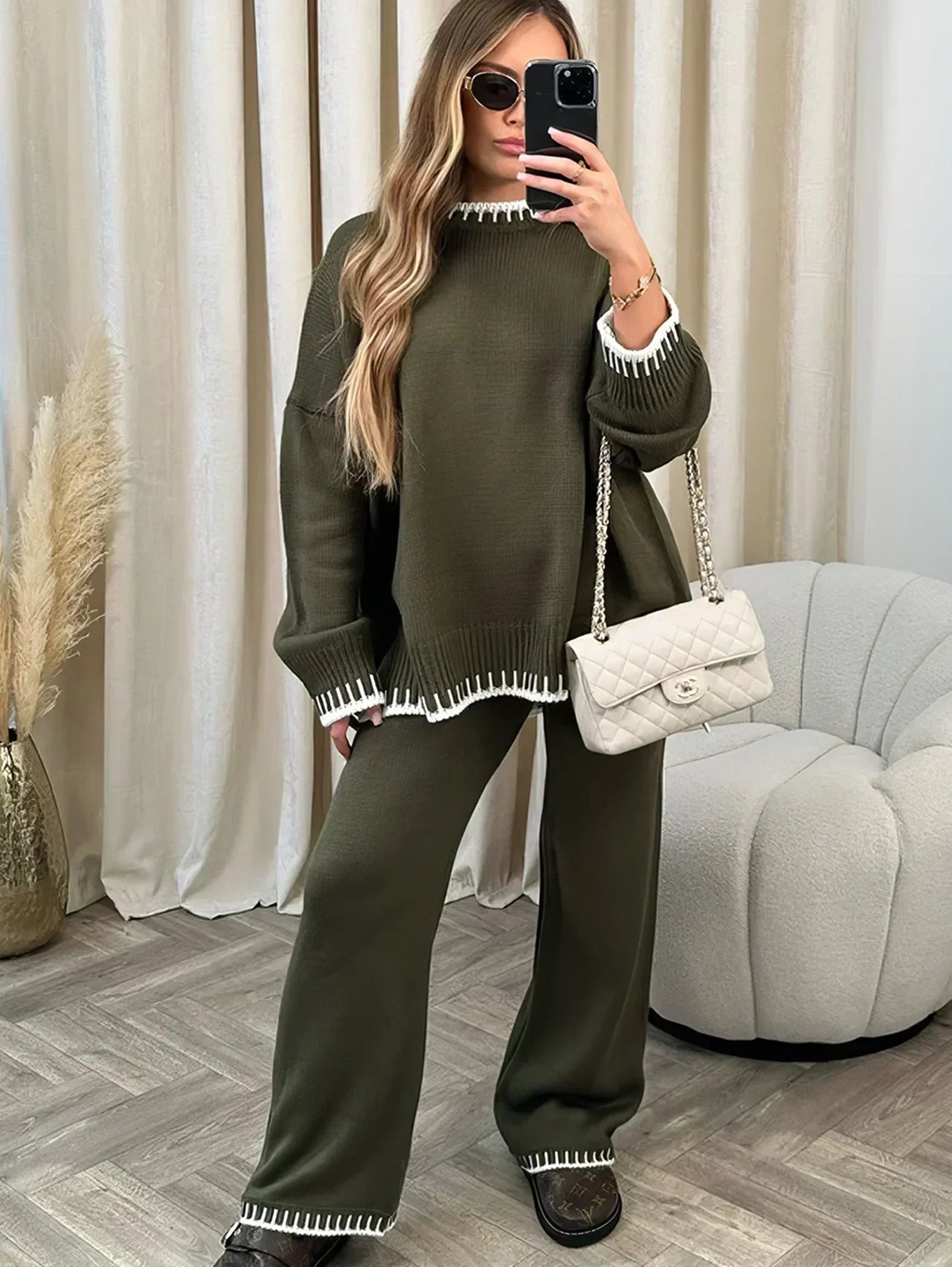 Femme prenant un selfie, habillée d’un ensemble lounge vert kaki avec finitions blanches contrastantes au col, poignets, ourlets et bas du pantalon. L’ensemble comprend un pull ample à manches longues et un pantalon large assorti. Look cosy, chic et confortable, accessoirisé d’un sac matelassé beige et de mocassins foncés.