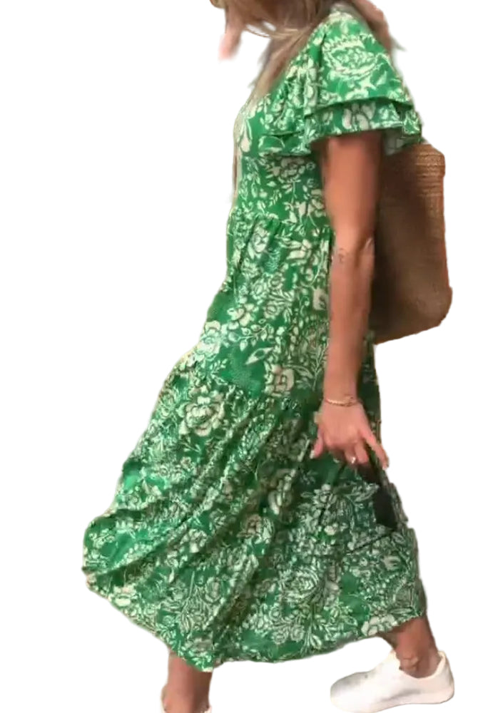 Femme marchant en robe verte à fleurs, associée à un sac en osier et des baskets blanches, dans un cadre extérieur lumineux.