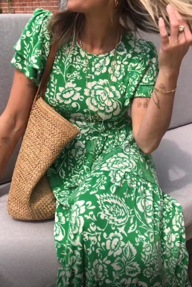 Femme assise sur un canapé extérieur, portant une robe verte à fleurs, un sac en osier, et des accessoires, tout en jouant avec ses cheveux.