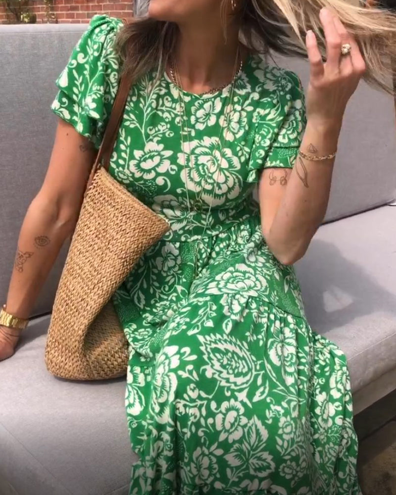 Femme assise sur un canapé extérieur, portant une robe verte à fleurs, un sac en osier, et des accessoires, tout en jouant avec ses cheveux.