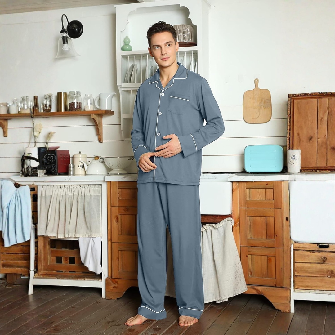 Homme debout dans une cuisine au style rustique, vêtu d’un pyjama bleu ardoise avec passepoils blancs et fermeture boutonnée. Ensemble confortable et raffiné, idéal pour les soirées cocooning ou les matinées détendues à la maison.