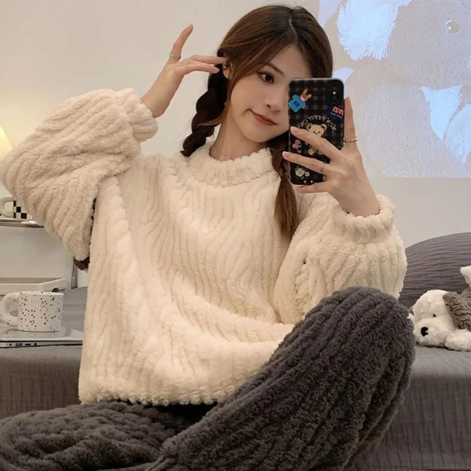 Pyjama femme confortable en peluche, composé d'un haut ample blanc et d'un pantalon côtelé gris foncé. La personne, assise sur un lit, prend un selfie en tenant son téléphone, affichant une expression détendue avec ses cheveux en tresses. L'ambiance cosy est accentuée par des peluches et une tasse sur la table à proximité.