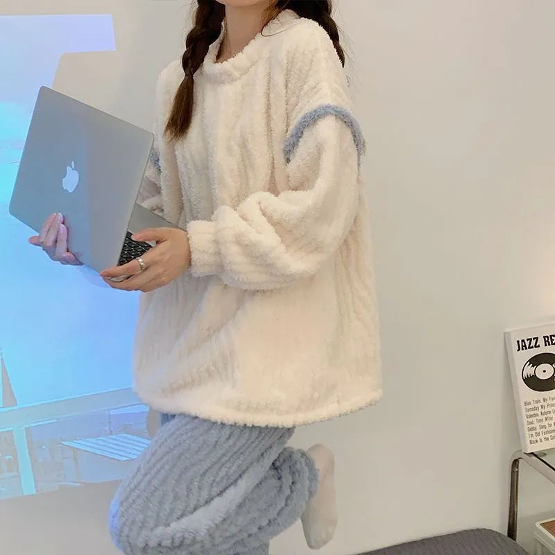 Pyjama femme en peluche avec un haut ample blanc et bleu clair, associé à un pantalon à texture côtelée. La personne tient un ordinateur portable tout en posant dans un décor minimaliste et lumineux. Elle affiche un sourire détendu et porte des vêtements confortables, parfaits pour une journée cosy à la maison.