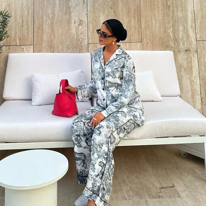 Pyjama femme Charline gris imprimé toile de Jouy, ensemble élégant et confortable deux pièces avec chemise boutonnée à col tailleur et pantalon long assorti, porté assise sur un canapé blanc, coupe fluide et chic.