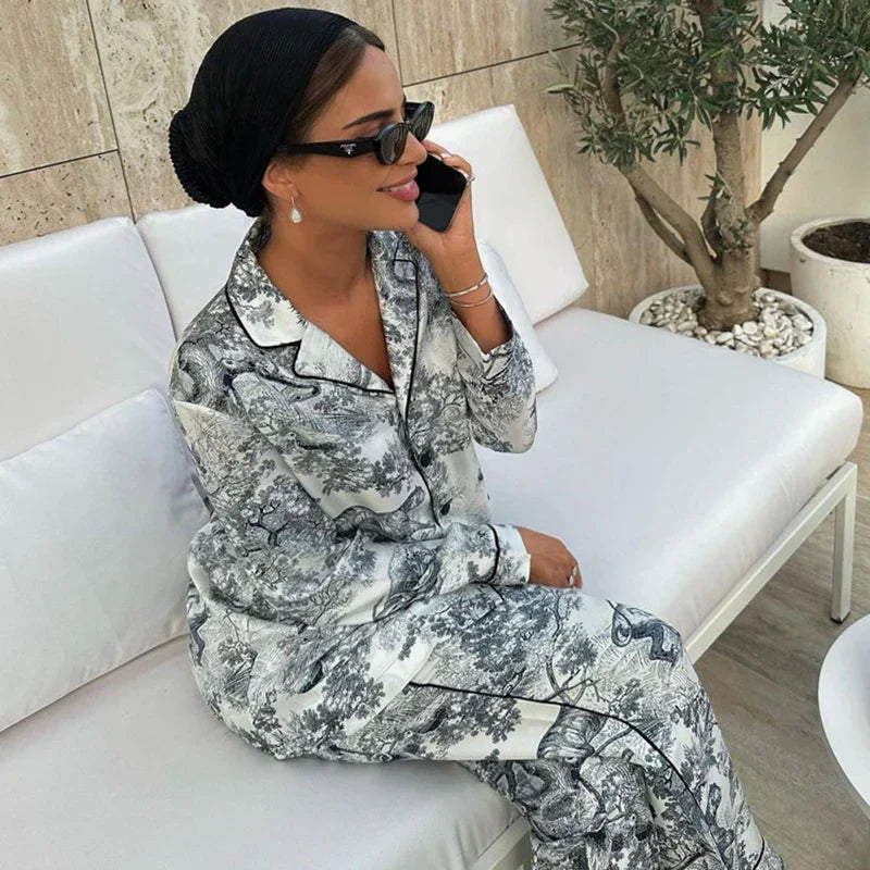 Pyjama femme Charline gris imprimé toile de Jouy, ensemble élégant et confortable deux pièces avec chemise boutonnée à col tailleur et pantalon long assorti, porté assise sur un canapé, finition passepoilée chic.