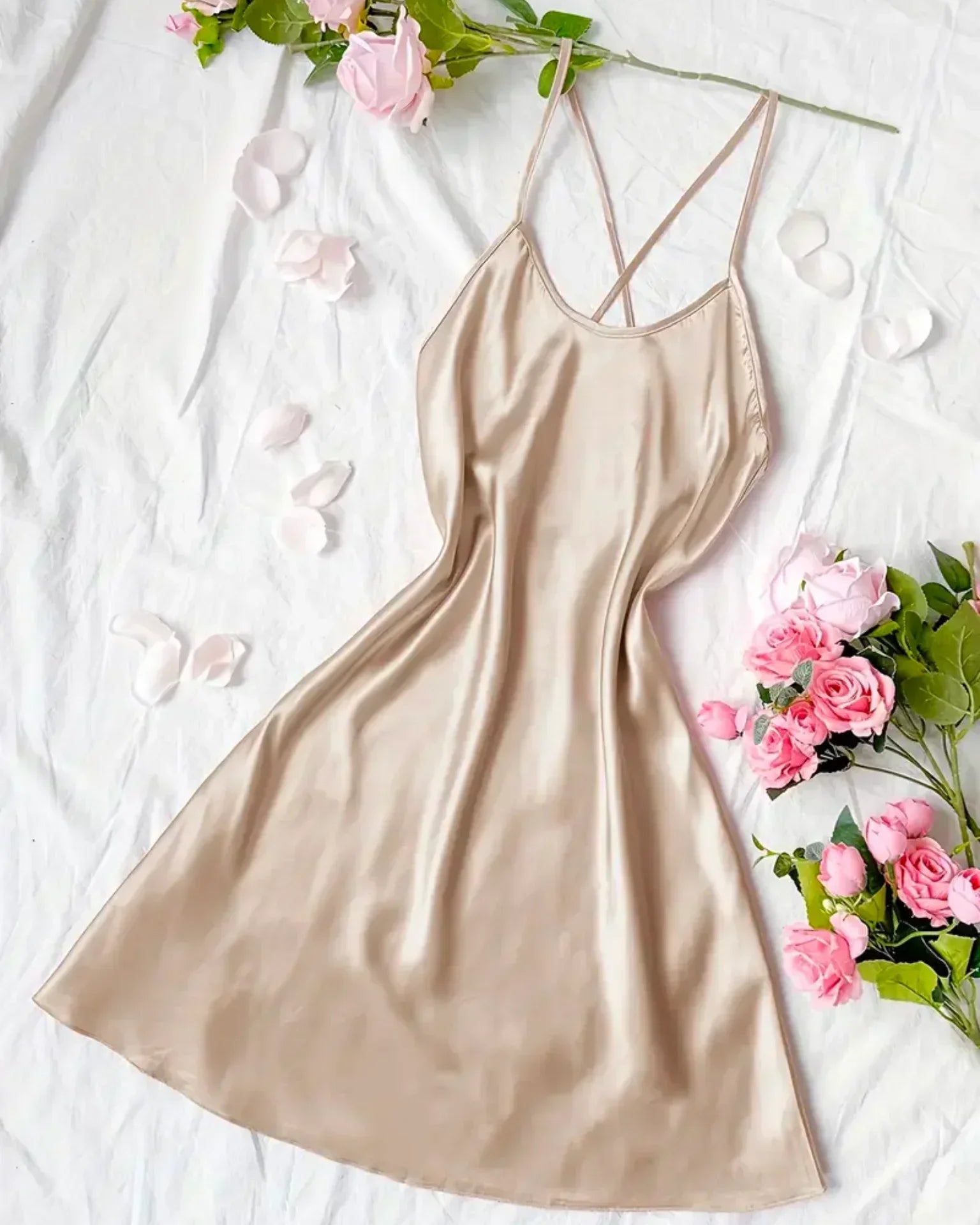 Nuisette femme Constance en satin couleur champagne, coupe courte et fluide avec fines bretelles croisées, présentée à plat sur un drap blanc avec roses et pétales, style élégant et confortable.