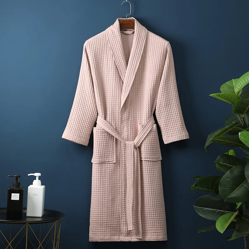 Peignoir Damien pour homme, modèle moelleux et confortable en tissu gaufré rose poudré, présenté suspendu devant un mur bleu avec accessoires de salle de bain en décor.