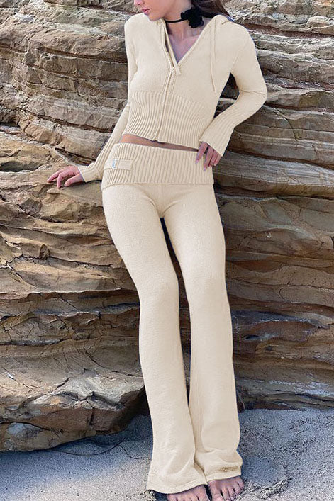 Femme posant sur une plage, vêtue de l’ensemble Dani en maille beige sable, composé d’un haut zippé à capuche avec manches longues et d’un pantalon taille basse évasé assorti. Tenue douce, féminine et élégante, parfaite pour un look confortable et tendance en bord de mer.