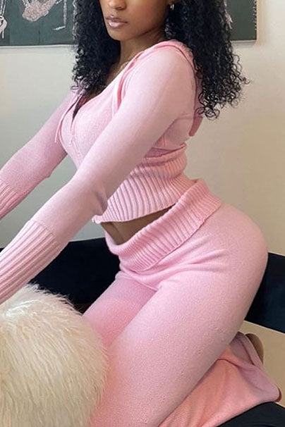 Femme assise portant l’ensemble Dani en maille rose pastel, composé d’un haut zippé à capuche et manches longues, et d’un pantalon taille haute assorti. Tenue douce, élégante et féminine, parfaite pour un look décontracté et tendance à la maison.