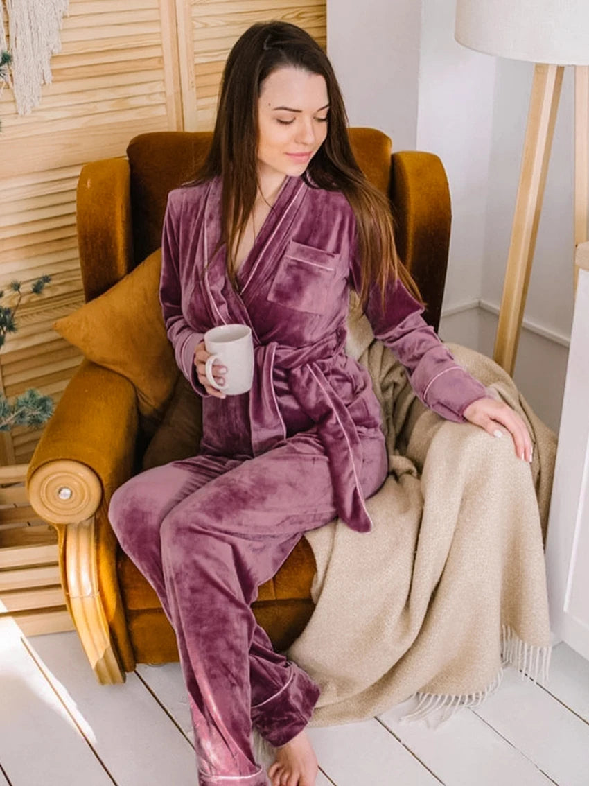 Femme assise dans un fauteuil, portant le pyjama Elysia en velours mauve avec veste croisée ceinturée et pantalon assorti. Ensemble chaud, doux et très confortable, parfait pour se détendre à la maison avec une boisson chaude.