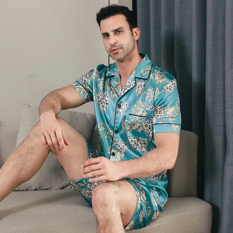 Homme assis portant le pyjama satin Georges pour homme, ensemble deux pièces avec chemise boutonnée à manches courtes et short assorti, imprimé décoratif vert turquoise, présenté en intérieur.
