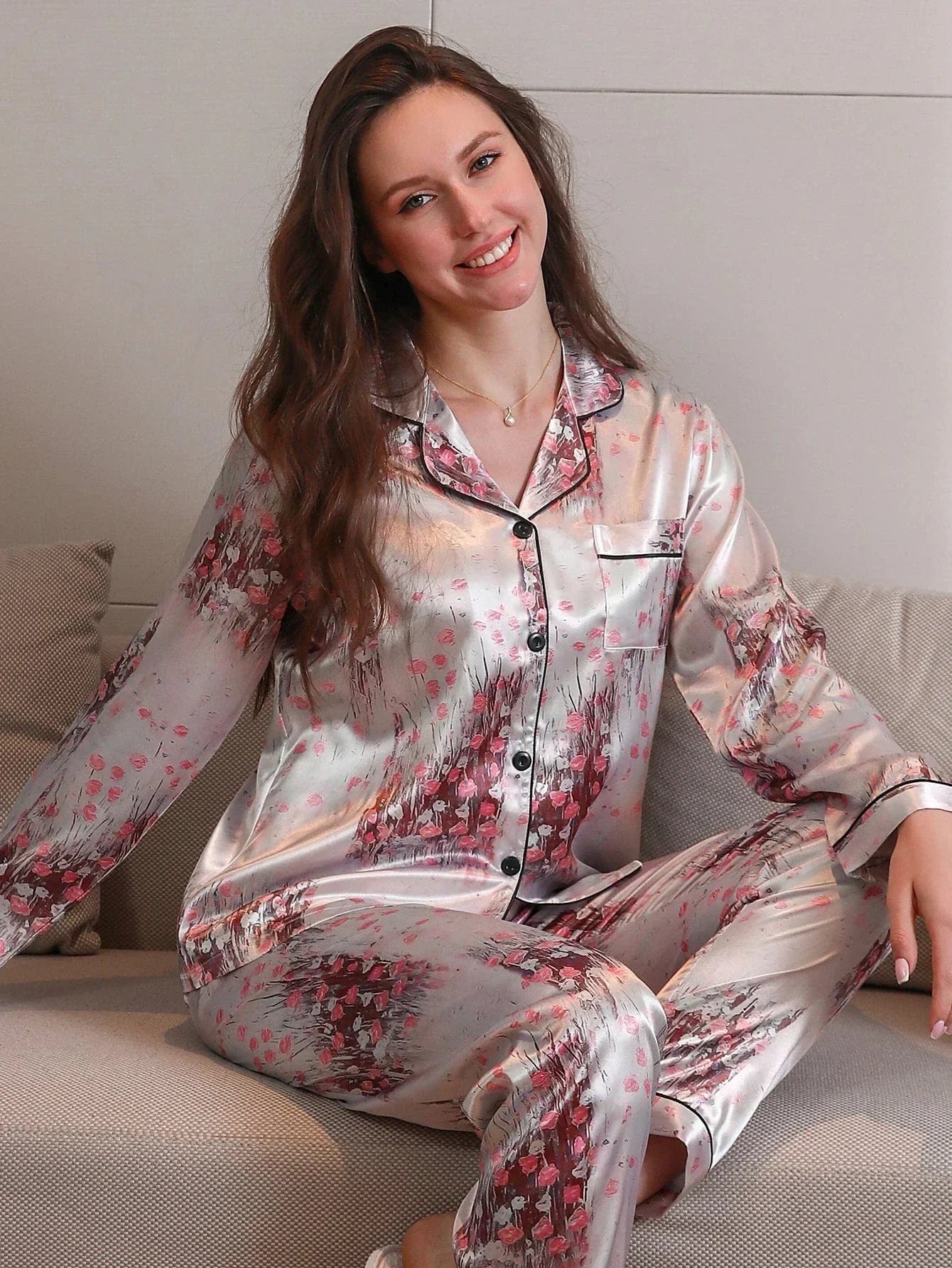 Femme assise et souriante sur un canapé, portant un pyjama long en satin gris perle orné de motifs floraux roses. Haut à manches longues boutonné avec col passepoilé noir, assorti à un pantalon fluide. Ensemble doux, confortable et raffiné, parfait pour des moments de détente avec style.