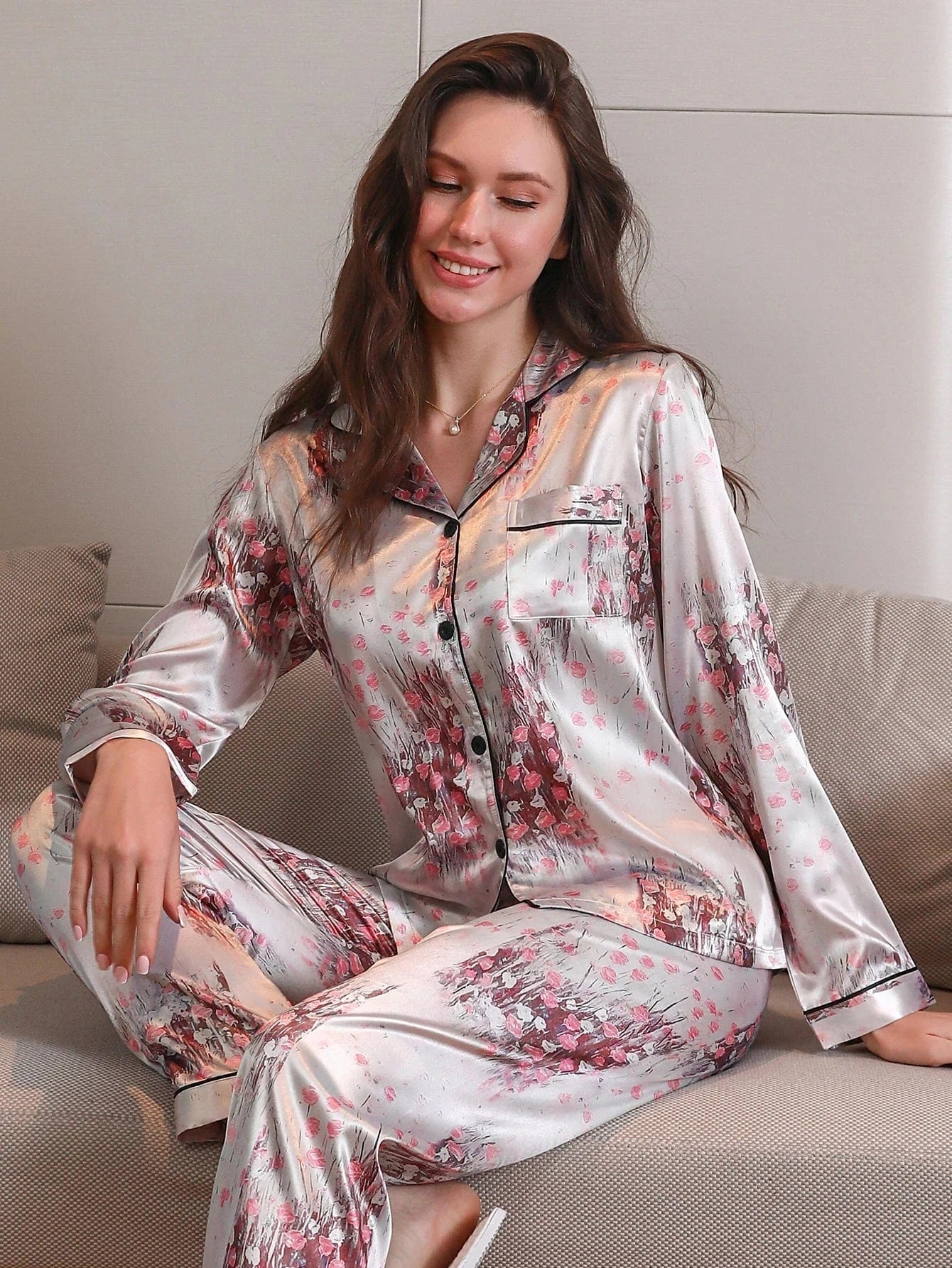 Femme assise en tailleur sur un canapé, portant un pyjama long en satin gris clair avec imprimés floraux roses. Haut boutonné à manches longues avec col passepoilé noir et poche poitrine, assorti à un pantalon fluide. Ensemble doux, élégant et confortable, idéal pour les soirées cocooning avec style.