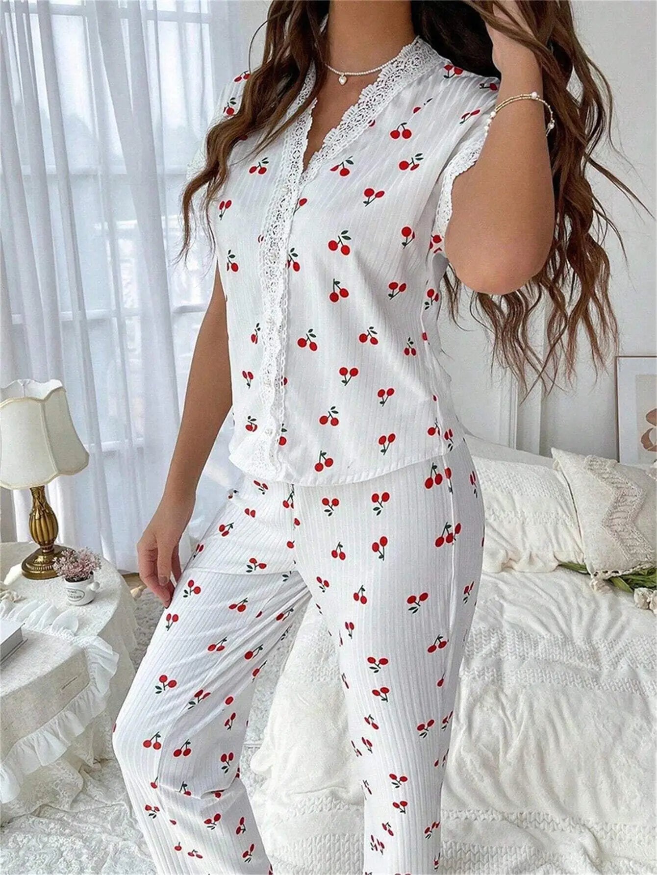 emme portant un pyjama léger et confortable, modèle "Léonie", en tissu blanc à rayures fines orné de motifs cerises rouges. Le haut à manches courtes est décoré de dentelle blanche sur le col et la boutonnière, assorti à un pantalon long avec finition en dentelle au bas. La scène se déroule dans une chambre lumineuse à la décoration élégante, créant une ambiance douce et reposante.