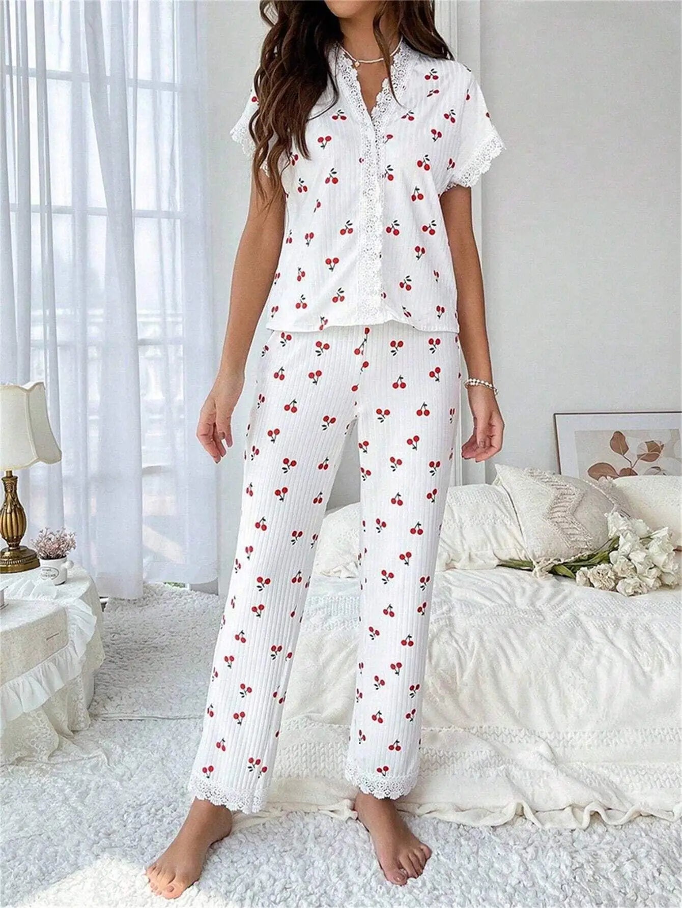 Femme debout dans une chambre lumineuse portant le pyjama "Léonie", un ensemble léger et confortable en tissu blanc à rayures verticales avec motifs de petites cerises rouges. Le haut à manches courtes est orné de dentelle délicate sur le col et les manches, assorti à un pantalon long avec finitions en dentelle aux chevilles. L’ambiance de la pièce est douce et cosy, idéale pour des moments de détente à la maison.