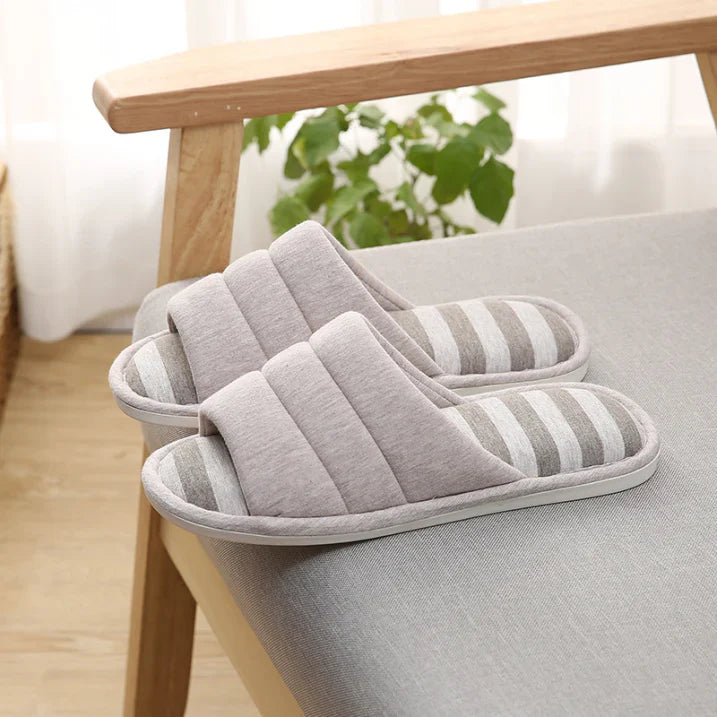 Paire de chaussons femme ouverts gris clair avec semelle à rayures grises et blanches, posés sur un fauteuil. Modèle doux et rembourré pour un confort cosy, avec semelle antidérapante idéale pour la maison.