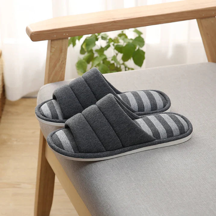 Paire de chaussons femme ouverts gris foncé avec semelle à rayures grises, posés sur un fauteuil. Chaussons doux et rembourrés pour un confort cosy, avec semelle antidérapante idéale pour la maison.
