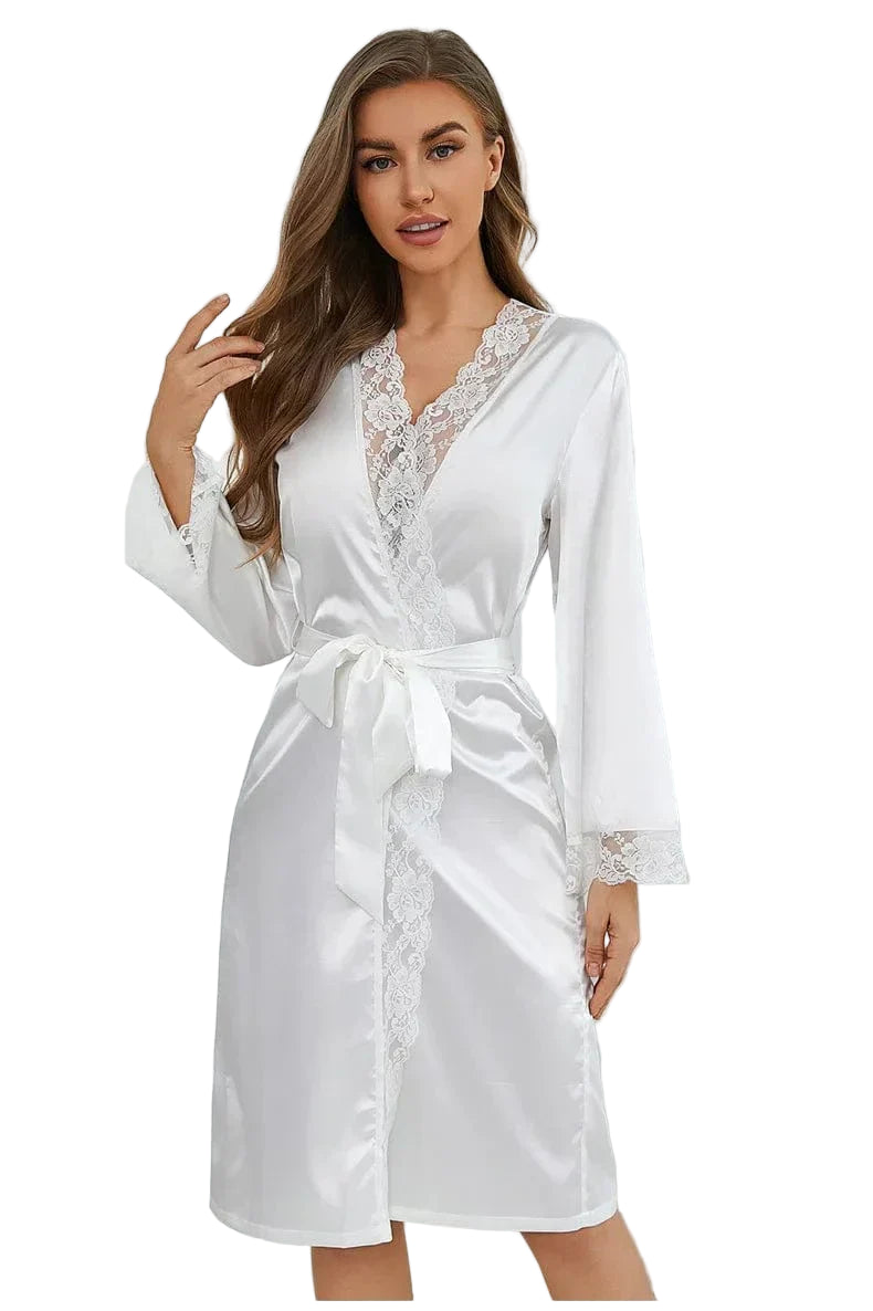 Femme portant un peignoir blanc en satin orné de délicates finitions en dentelle sur le col, les poignets et l’ouverture. Ceinture nouée à la taille pour une silhouette féminine et raffinée. Modèle élégant, idéal pour la mariée ou pour une touche glamour au quotidien.