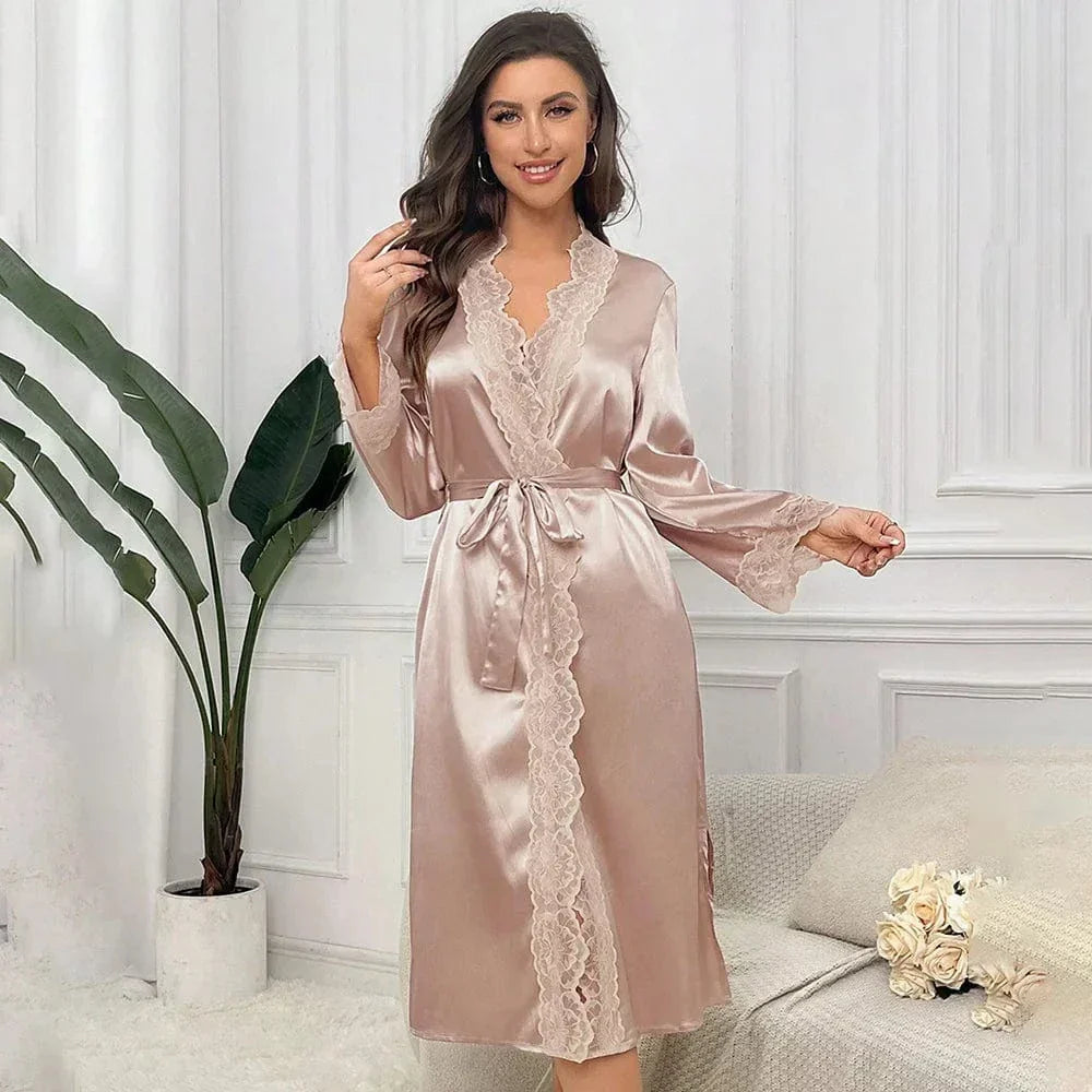 Femme souriante portant le peignoir Mélodie en satin champagne avec finition dentelle délicate. Le modèle mi-long présente une ceinture à nouer à la taille et des manches longues, pour un look à la fois élégant et confortable, parfait pour la maison. Le décor raffiné ajoute une touche chaleureuse à la scène.