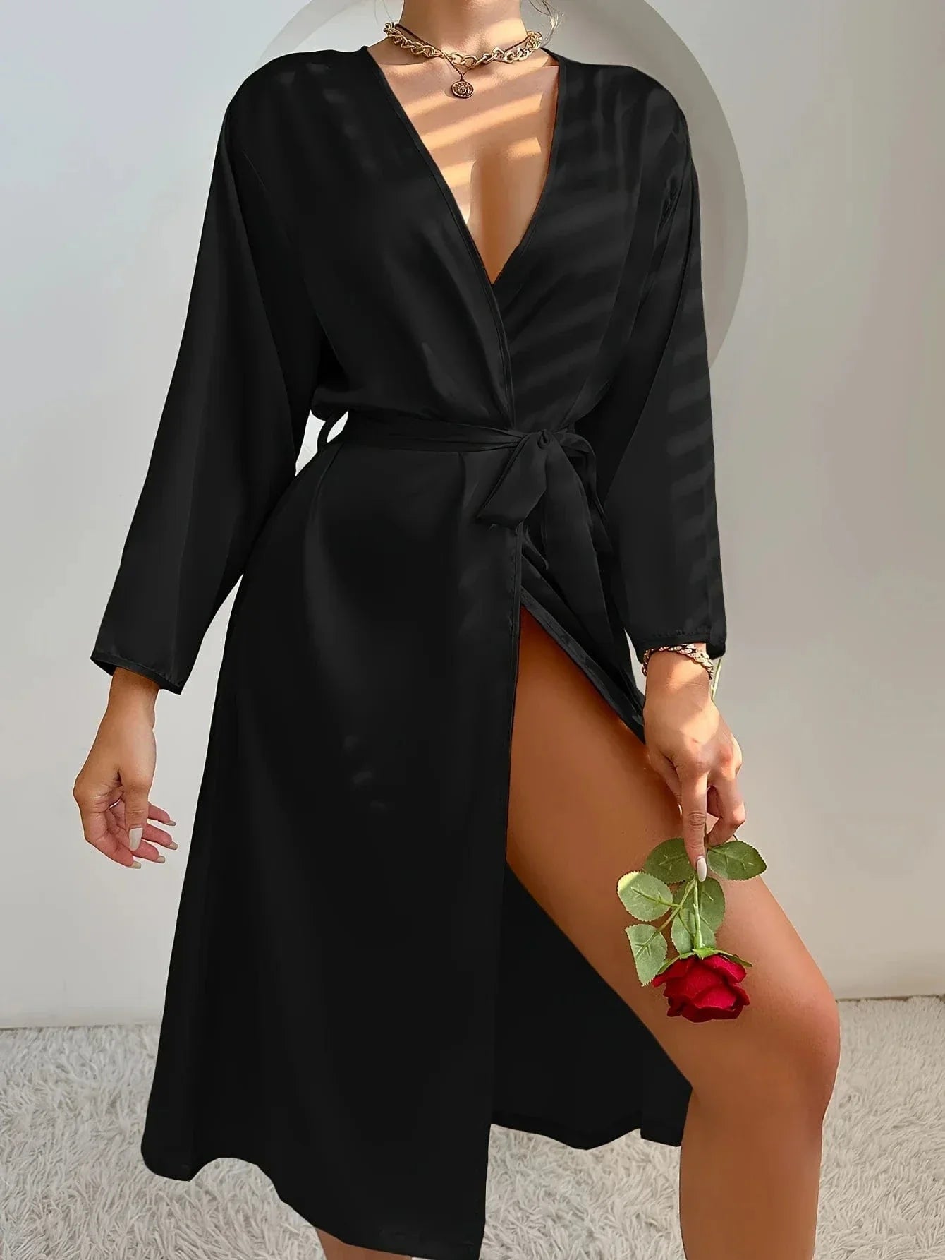 Femme debout, tenant une rose rouge, vêtue d’un peignoir noir satiné à col croisé et ceinture à nouer. Coupe fluide et fendue, pour une allure élégante et sensuelle. Parfait pour des moments de détente raffinés ou un look romantique à la maison.