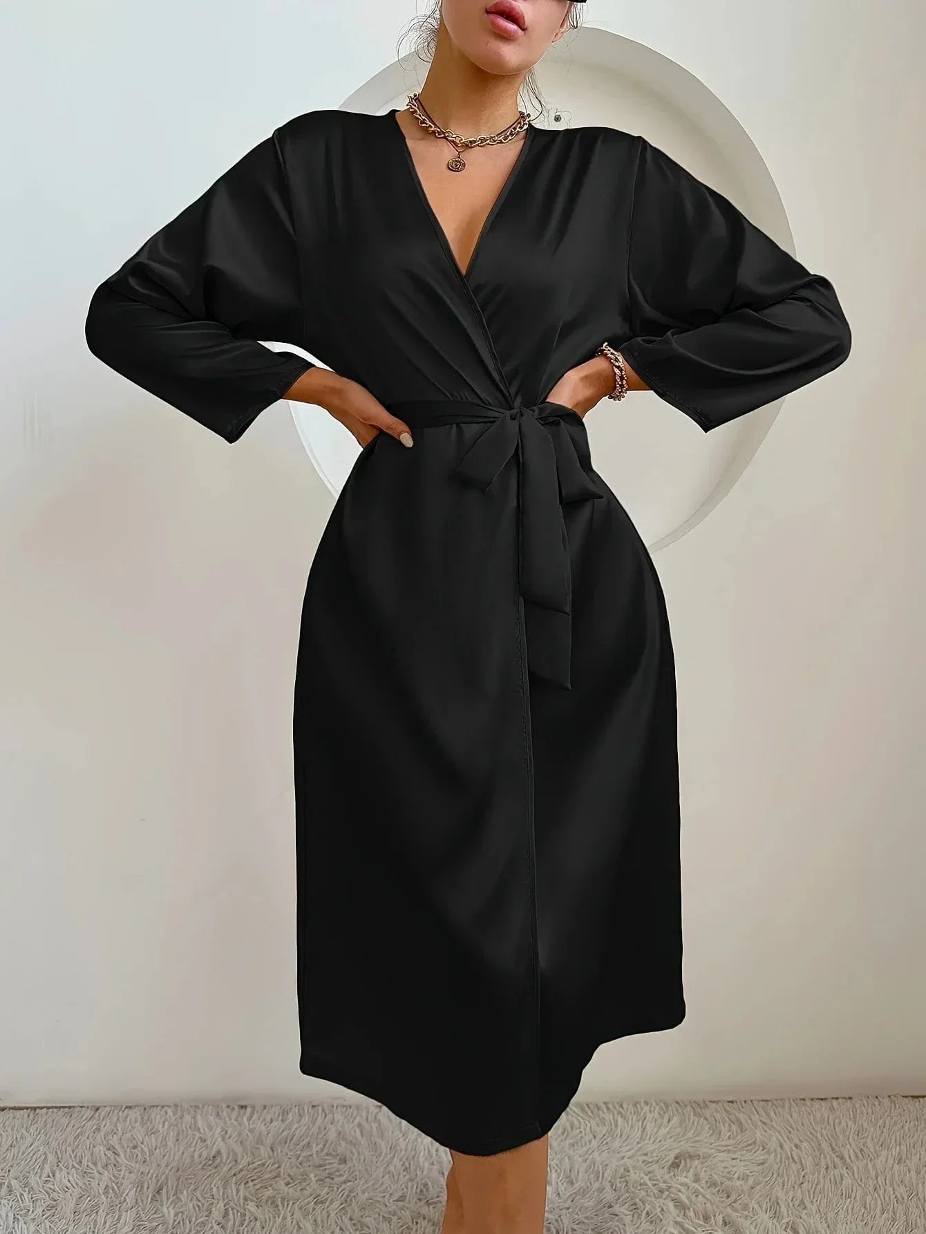 Femme debout, portant un peignoir noir satiné à manches longues, coupe midi et ceinture nouée à la taille. Modèle sobre, fluide et élégant, idéal pour des moments de détente confortables avec une allure chic et raffinée.