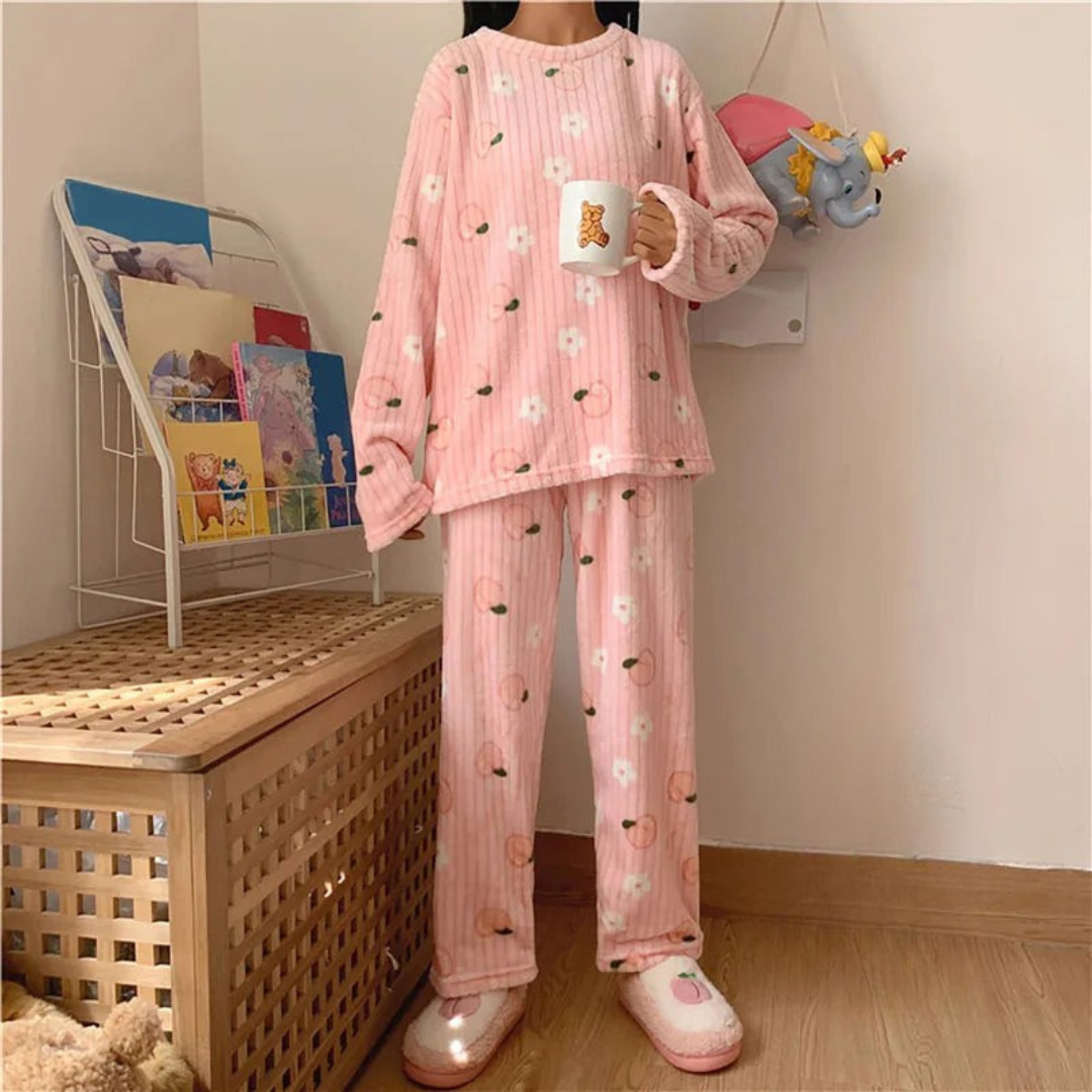 Pyjama femme rose en peluche avec des motifs de pêches et de fleurs blanches. La personne porte un haut à manches longues et un pantalon assorti, tenant une tasse décorée d'un ours. Elle porte également des chaussons mignons, créant une ambiance cosy et confortable, parfaite pour se détendre à la maison.