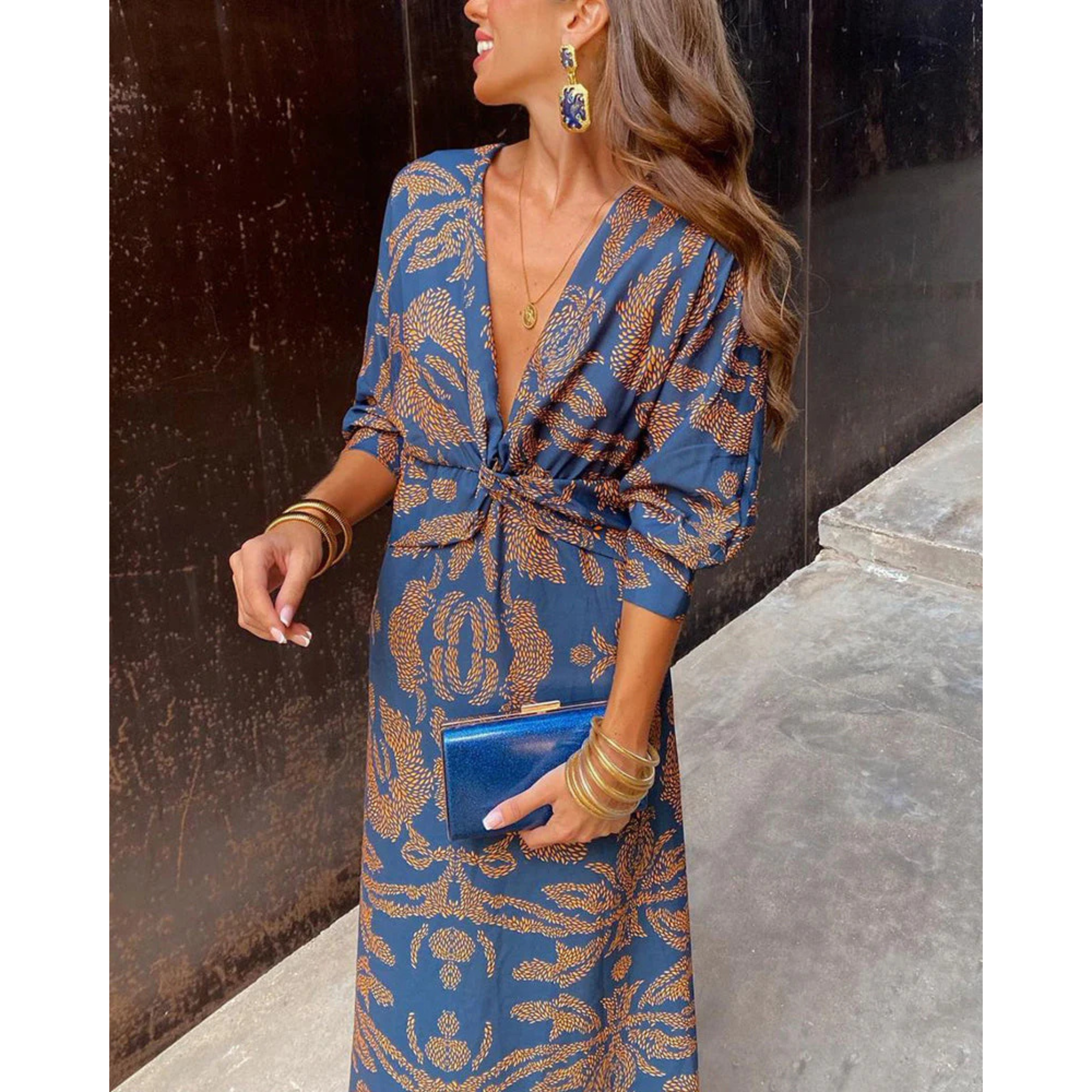 Robe portefeuille bleue à motifs dorés, décolleté en V et manches 3/4, nouée à la taille, avec bijoux dorés et pochette bleue.