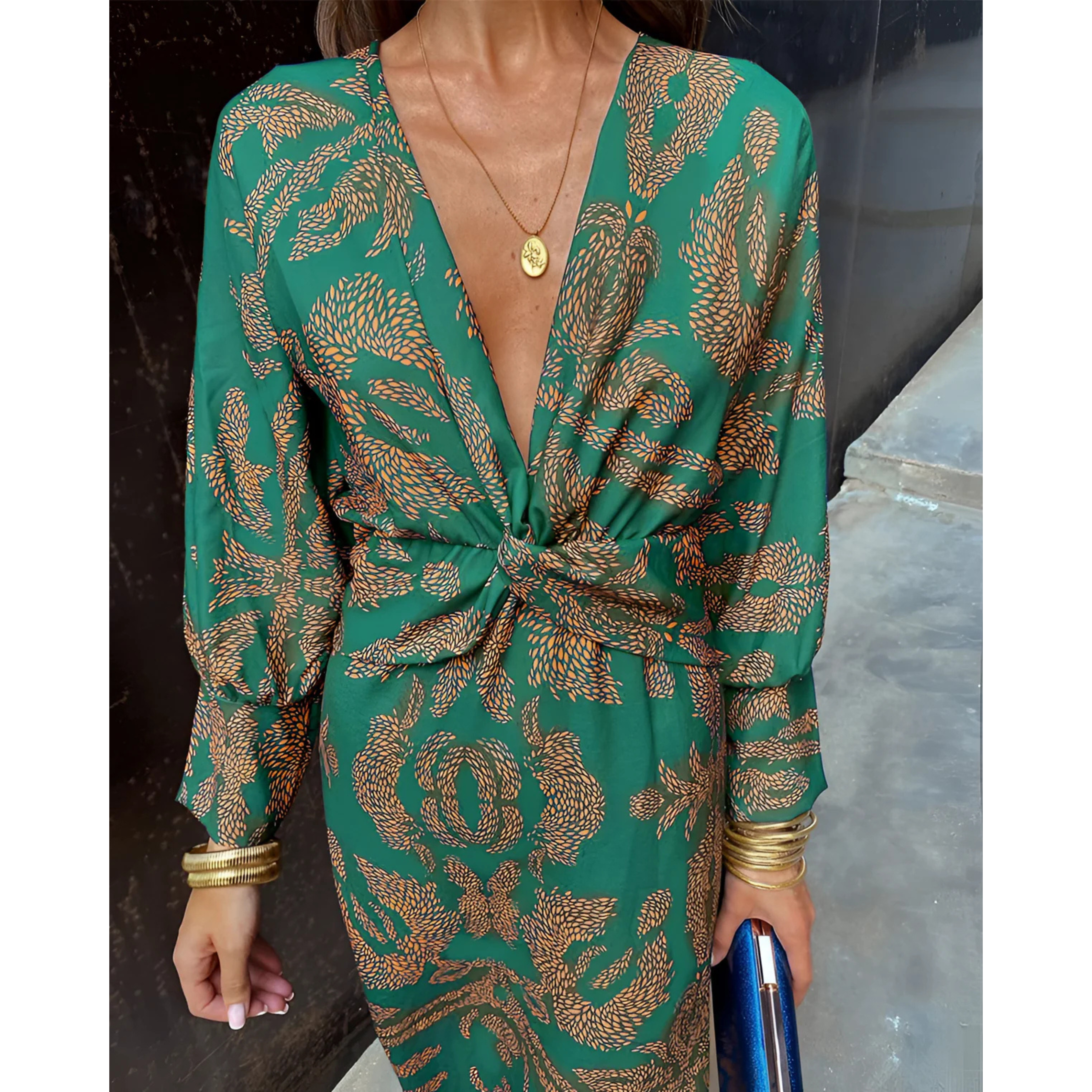 Robe portefeuille verte à motifs dorés, décolleté en V profond et taille nouée, manches longues, portée avec bijoux dorés et pochette bleue.