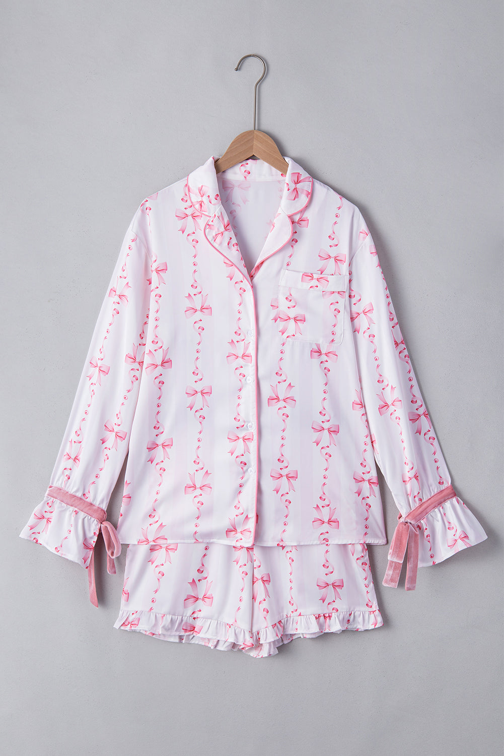 Pyjama femme Pascale blanc à motifs nœuds roses, ensemble deux pièces suspendu sur cintre avec chemise boutonnée à col et poche poitrine, manches longues avec poignets volantés et liens à nouer, short assorti à ourlet volanté.