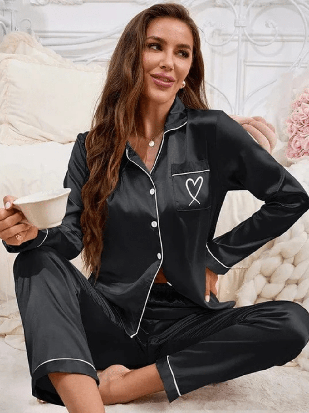 Femme assise sur un lit, portant un pyjama noir satiné à boutons avec passepoils blancs, col à revers et poche poitrine ornée d’un motif cœur. Ensemble élégant, doux et agréable à porter, parfait pour des moments de détente à la maison.