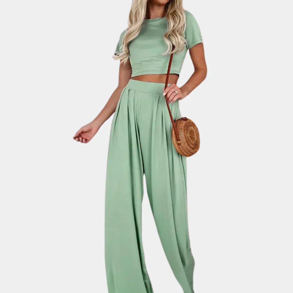 Femme portant l’ensemble Rosalind vert sauge, composé d’un crop top à manches courtes et d’un pantalon palazzo fluide taille haute à plis. Elle accessoirise la tenue avec un sac rond en osier. Look estival, léger et confortable, parfait pour une allure chic et décontractée.