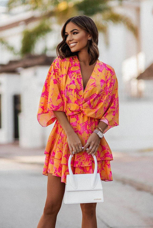 Femme souriante portant une robe mini fleurie aux couleurs vives de rose et orange, avec un design fluide et léger. Idéale pour l'été, la robe offre confort et élégance avec des volants et une coupe flatteuse.