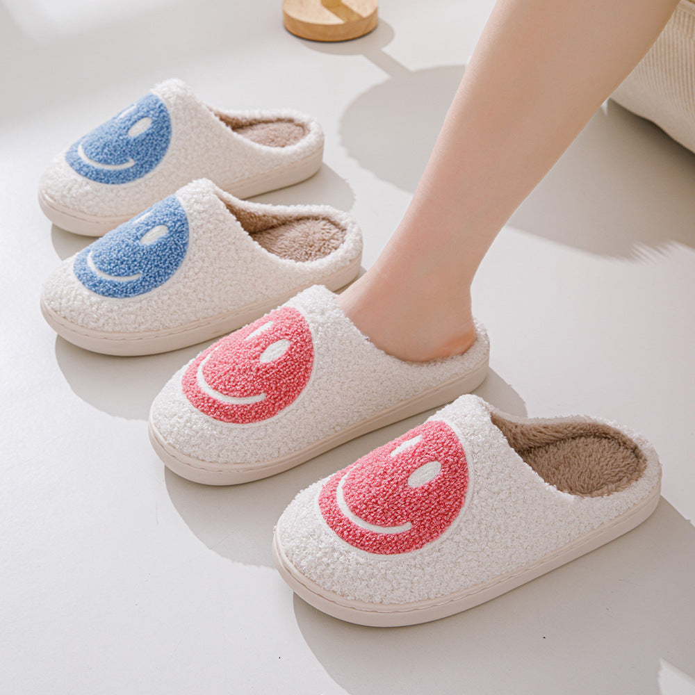 Chaussons unisexe Sasha blancs en matière pelucheuse avec smiley rose, portés aux pieds, avec modèles smiley bleu en arrière-plan, intérieur chaud et cosy pour la maison.