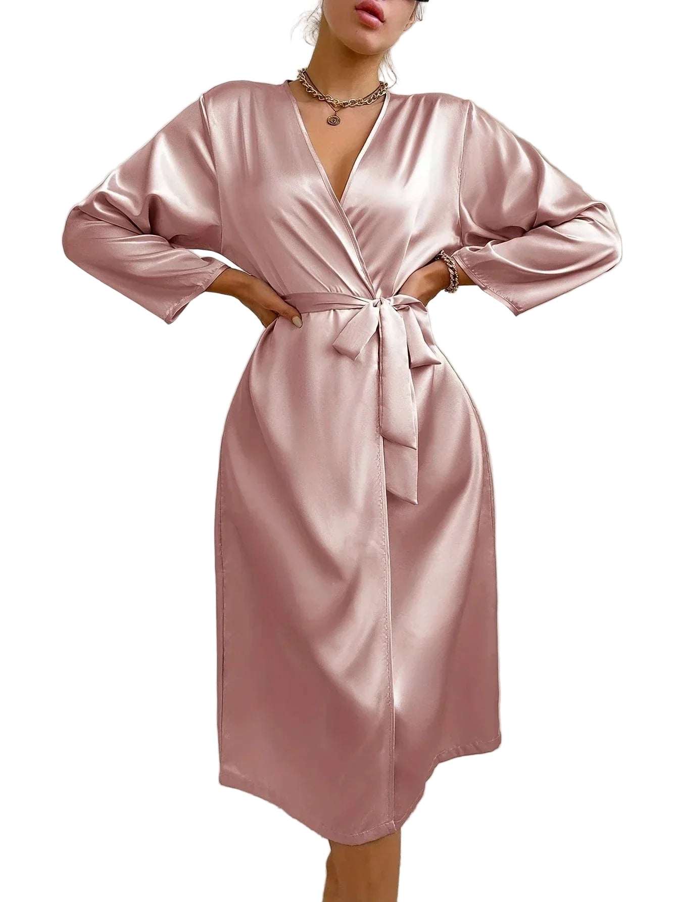 Femme debout, portant un peignoir satiné rose poudré à col croisé, manches longues et ceinture nouée à la taille. Modèle mi-long, fluide et raffiné, parfait pour une allure chic à la maison ou en tenue de nuit élégante.
