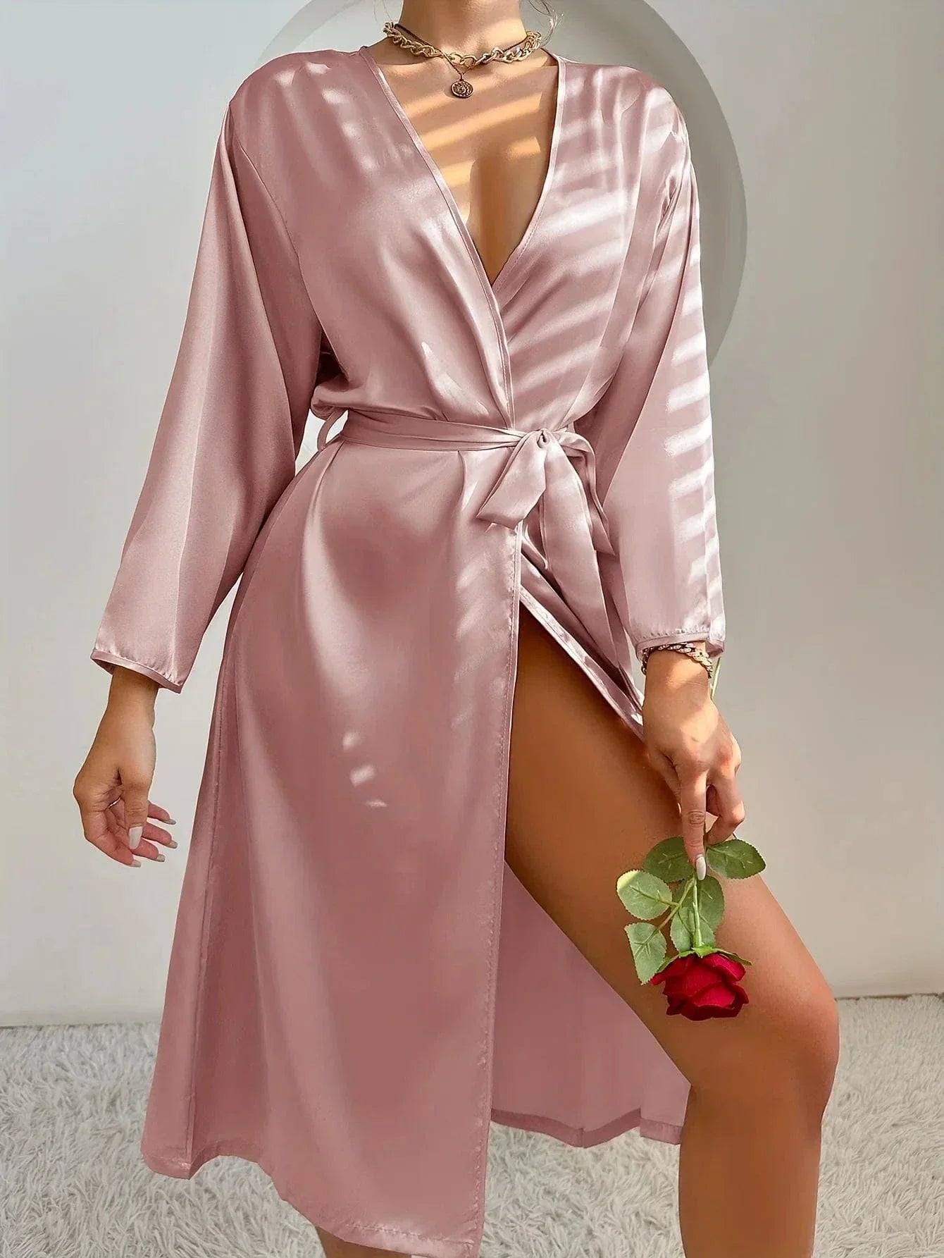 Femme debout tenant une rose rouge, vêtue d’un peignoir satiné rose poudré à col croisé, ceinture à nouer et fente élégante sur la jambe. Modèle mi-long fluide et raffiné, idéal pour une tenue de nuit chic ou un moment de détente avec style.
